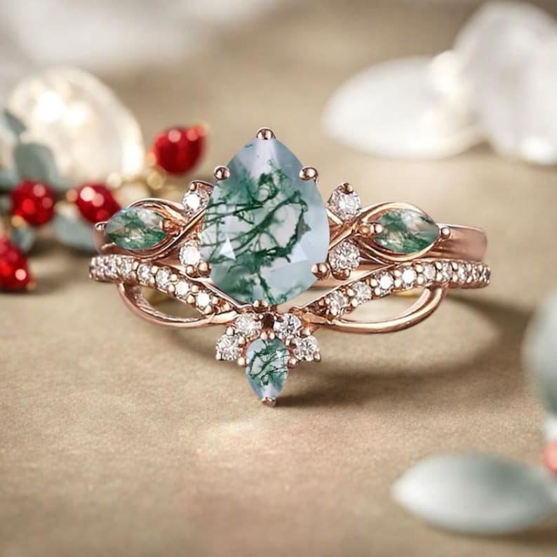 May include: Rose gold ring set featuring a pear-shaped moss agate gemstone with green veining. The ring is accented with small diamonds and two marquise-cut green gemstones. A second band with small diamonds completes the set.