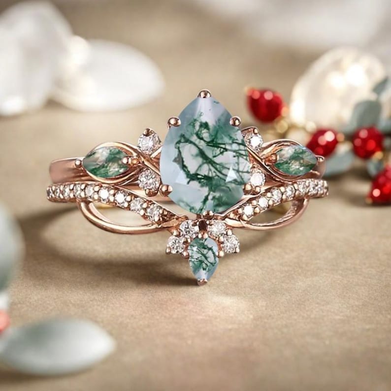 May include: A rose gold ring set featuring a pear-shaped moss agate gemstone with green inclusions. The ring is accented with small diamonds and additional green gemstones. The band has a delicate, intertwined design.