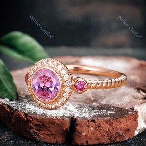 May include: A rose gold engagement ring featuring a pink gemstone encircled by a halo of smaller diamonds. Two smaller pink gemstones are set on the band.