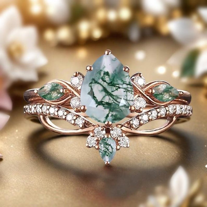 May include: A rose gold ring set featuring a pear-shaped moss agate gemstone with green inclusions. The engagement ring is accented with small diamonds and green gemstones. The wedding band is a delicate design with small diamonds.