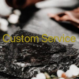 May include: A close-up shot of a dark, textured surface with the words "Custom Service" in yellow. The background is blurred, with hints of white flowers and a hand. The overall aesthetic is natural and organic.