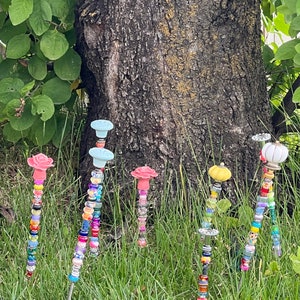 May include: Six colorful beaded garden stakes with different colored ceramic tops. The tops are shaped like flowers and a pumpkin. The stakes are in a grassy area in front of a tree.