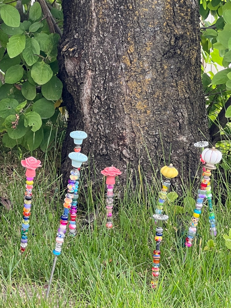 May include: Six colorful beaded garden stakes with different colored ceramic tops. The tops are shaped like flowers and a pumpkin. The stakes are in a grassy area in front of a tree.