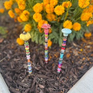 Può includere: Tre paletti da giardino colorati con cime in ceramica di diversi colori e perline colorate. I paletti sono in un letto di pacciame marrone.