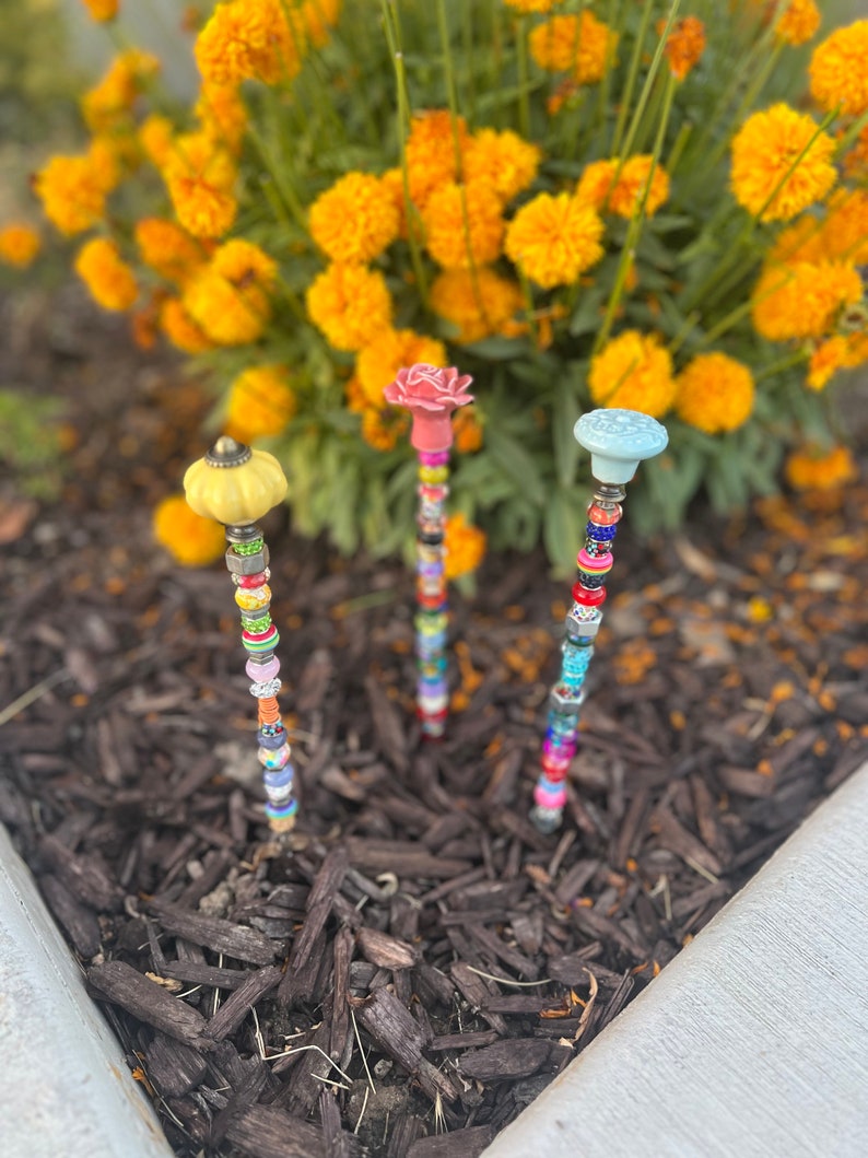 May include: Three colorful garden stakes with different colored ceramic tops and colorful beads. The stakes are in a bed of brown mulch.