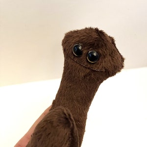 May include: A brown plush toy with a long neck and a round head. The toy has two large black eyes and a small mouth.