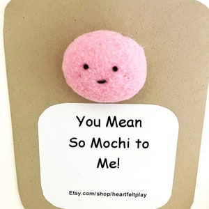 May include: A pink felt mochi character with a smiling face is centered on a brown card. The card has a white label with the text "You Mean So Mochi to Me!"