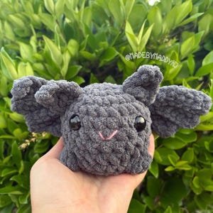 May include: A handmade gray crochet bat with large, textured wings and black safety eyes. The bat has a small pink stitched smile. The image includes the text "@madebymaddy".