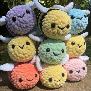 May include: A stack of nine crocheted bee plushies in various colors, including purple, yellow, black, light blue, green, pink, and orange. Each bee has a smiling face with black eyes and a white crocheted wing.