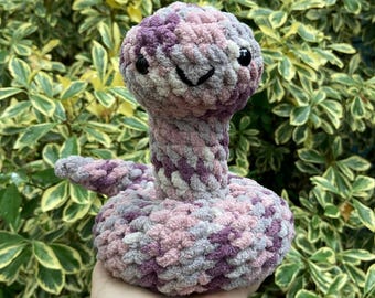 May include: A crocheted plush toy shaped like a snake with a purple, pink, and gray color scheme. The snake has a smiling face with black eyes.
