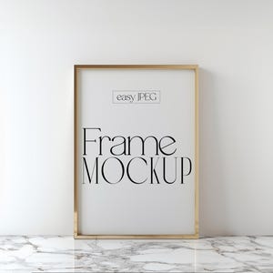 Puede incluir: Una maqueta de marco dorado con el texto "Frame Mockup" y "easy JPEG" sobre un fondo blanco. El marco está sobre una superficie de mármol blanco.