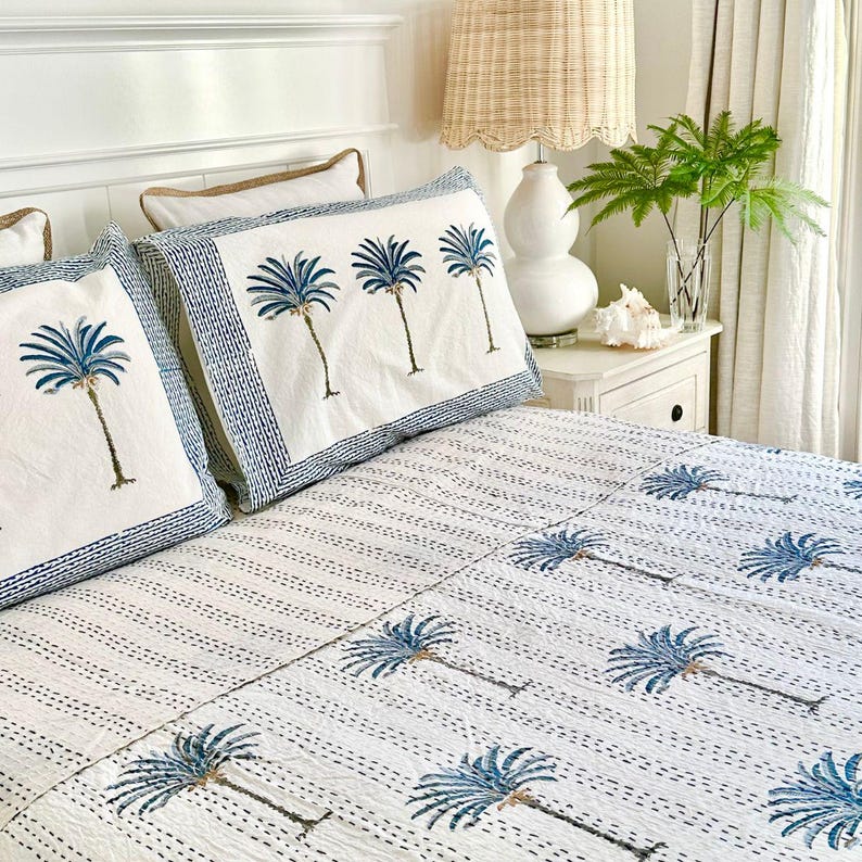 May include: White bed with blue and white patterned bedding featuring a palm tree design. The bedding is made of cotton and has a quilted texture. The bed is made up with two pillows, one with a solid white cover and the other with a blue and white patterned cover. A lamp with a wicker shade is on a white nightstand next to the bed.