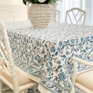 May include: A white tablecloth with a blue floral pattern. The tablecloth is laid on a table with white chairs.
