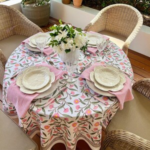 May include: A round table set for dining with a floral tablecloth featuring pink flowers and green leaves. The table is set with cream-colored plates, pink striped napkins, and clear glassware. Wicker chairs surround the table, and a vase of white flowers sits in the center.