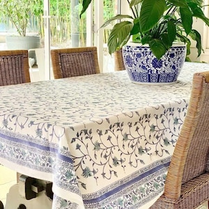 May include: A white tablecloth with a blue floral pattern covers a wooden table. The tablecloth has a blue border with a repeating floral design. Wicker chairs surround the table, and a blue and white patterned planter with a green plant sits on the table.