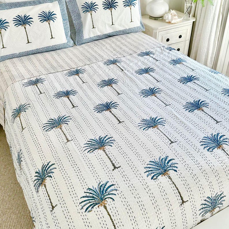 May include: White cotton quilt with a blue palm tree pattern. The quilt is on a bed with white sheets and two blue and white patterned pillowcases.