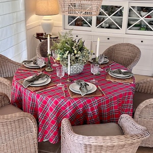 Peut inclure: Une table à manger ronde recouverte d'une nappe à carreaux rouges, verts et blancs, entourée de quatre chaises en osier tressé. La table est dressée avec des assiettes blanches, des verres texturés, des serviettes vert olive et un centre de table.