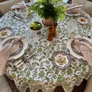 May include: A round table set for dining, featuring a white tablecloth with a green floral pattern. The table is set with plates, napkins, glasses, and silverware. A bowl of limes and a potted plant are in the center.
