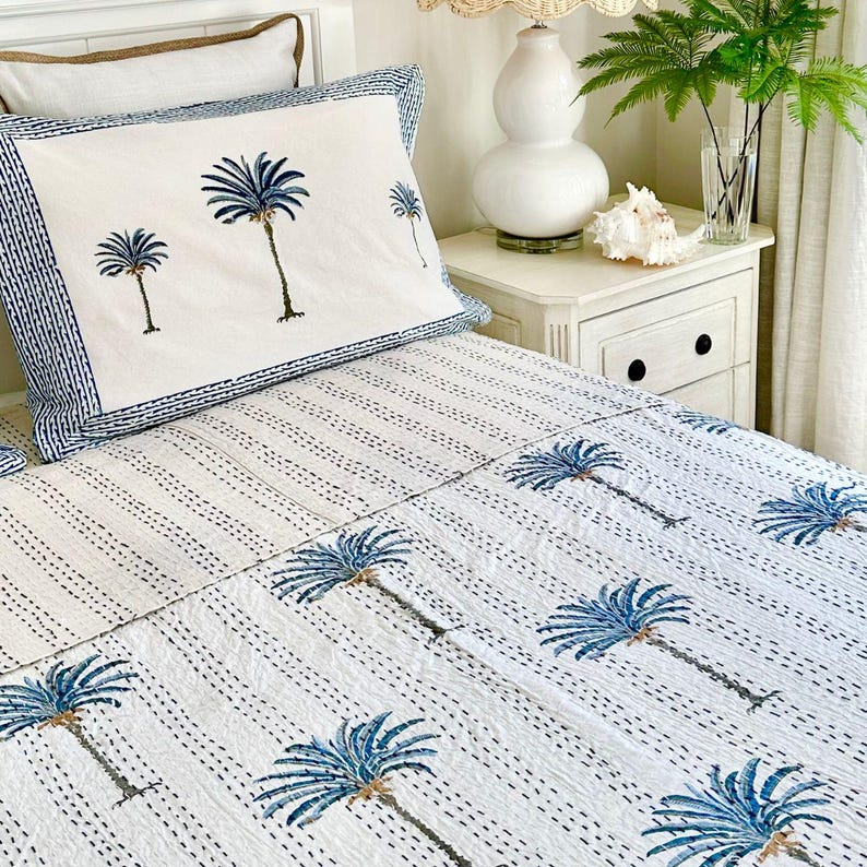 May include: White cotton bedding with a blue palm tree print. The bedding is on a bed with a white headboard and a white nightstand.