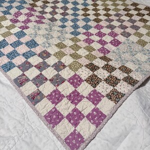 May include: A handmade quilt featuring a checkerboard pattern in shades of purple, blue, green, and white. The quilt has a floral print in some of the squares and a lavender border. The quilt is made of soft fabric.