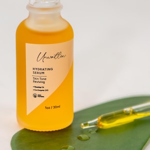 Puede incluir: Un frasco de vidrio de suero hidratante Unwritten, un suero revitalizante para el tono de la piel con un gotero, sobre una hoja verde. El frasco est&aacute; etiquetado con el nombre del producto, los ingredientes y el tama&ntilde;o: 1 oz / 30 ml.