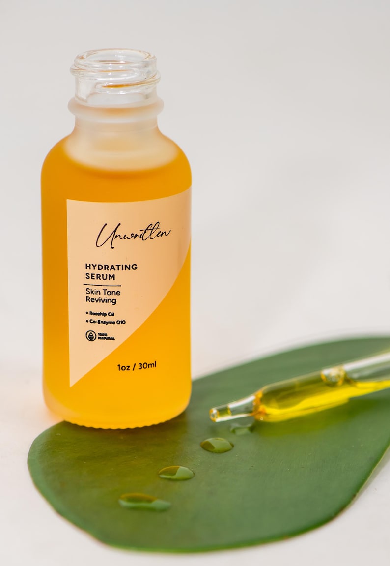 Puede incluir: Un frasco de vidrio de suero hidratante Unwritten, un suero revitalizante para el tono de la piel con un gotero, sobre una hoja verde. El frasco est&aacute; etiquetado con el nombre del producto, los ingredientes y el tama&ntilde;o: 1 oz / 30 ml.