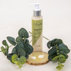 Puede incluir: Una botella blanca de limpiador de brillo c&iacute;trico con un dispensador de bomba. La botella est&aacute; etiquetada como "Citrus Glow Cleanser" y tiene un dise&ntilde;o verde y blanco. La botella est&aacute; sobre un posavasos de madera con hojas verdes alrededor.