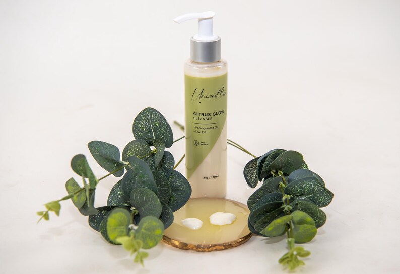 Puede incluir: Una botella blanca de limpiador de brillo c&iacute;trico con un dispensador de bomba. La botella est&aacute; etiquetada como "Citrus Glow Cleanser" y tiene un dise&ntilde;o verde y blanco. La botella est&aacute; sobre un posavasos de madera con hojas verdes alrededor.