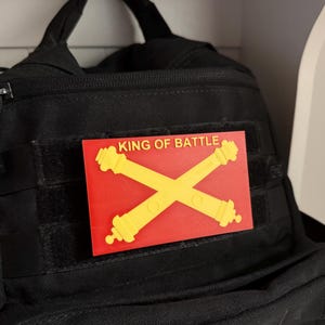 Op de afbeelding: Een rood en geel patch met de tekst "KING OF BATTLE" en een ontwerp met gekruiste kanonnen.