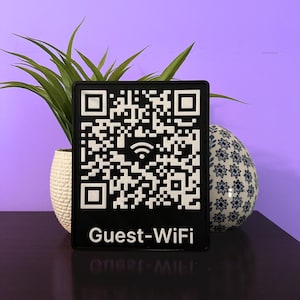 May include: A black rectangular sign with a white QR code and the words "Guest-WiFi" in white. The sign is displayed in front of a purple background, with a potted plant and a decorative egg-shaped object.