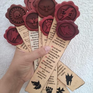 Könnte beinhalten: Eine Sammlung handgefertigter Lesezeichen mit geprägten roten Wachssiegeln. Jedes Lesezeichen zeigt ein einzigartiges Symbol und Text mit einem Vintage-Pergamentpapier-Aussehen. Die Siegel zeigen Schädel-, Adler- und Wolfsdesigns. Die Lesezeichen werden in einer Hand gehalten.
