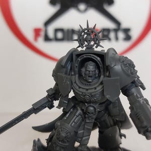 May include: A gray plastic miniature of a space marine with a sword and a helmet with a spiked crown. The miniature is unpainted.