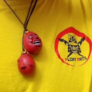 May include: A yellow t-shirt with a red circle logo featuring a black samurai warrior and the text "FLOIN ARTS". A brown cord with two red ceramic pendants with painted faces hangs from the neck of the shirt.