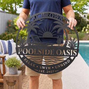 Op de afbeelding: Een rond zwart metalen bord met een uitgesneden ontwerp van een ligstoel, palmbomen en een zon. De tekst luidt THE BROWNS, POOLSIDE OASIS, RELAX, SWIM, UNWIND en EST. 2027. Het bord heeft een decoratieve rand en een matte afwerking.