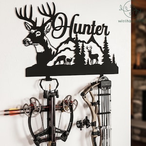 Op de afbeelding: Zwarte metalen wandkunst met een herten silhouet en het woord "Hunter". Het kunstwerk is gemonteerd boven een kruisboog en een boog, wat wijst op een jacht thema. De achtergrond heeft een berg- en bosontwerp.