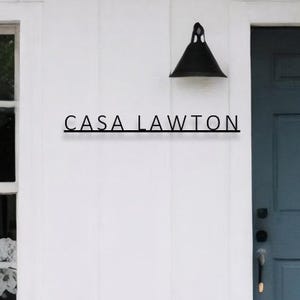 Könnte beinhalten: Ein schwarzes Metallschild mit dem Text "CASA LAWTON" an einer weißen Wand montiert. Eine schwarze Leuchte ist über dem Schild montiert.