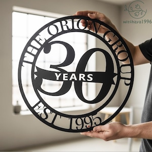 May include: A circular black metal sign featuring the text "THE ORION GROUP", "30 YEARS", and "EST 1995". The sign is held up by a person. The sign is a commemorative piece.