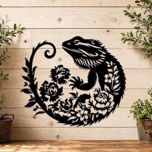 Puede incluir: Una pieza de arte mural de metal negro que presenta la silueta de un lagarto dragón barbudo integrado en un diseño circular. El lagarto está rodeado por intrincados recortes de flores y enredaderas, creando un detallado motivo floral.