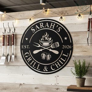 Pode incluir: Placa circular de metal preto com o texto "Sarah's Grill & Chill" e o ano de 2026. A placa apresenta utensílios de grelhar cruzados, uma tigela com chamas e duas canecas de cerveja. Luzes penduradas acima.