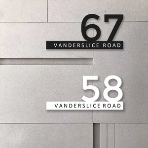 Könnte beinhalten: Zwei Hausnummern an einer grauen Betonwand. Die obere Nummer ist 67 und schwarz. Die untere Nummer ist 58 und weiß. Beide Nummern haben den Text "Vanderslice Road" darunter.