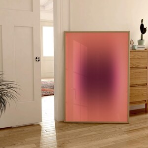 May include: A framed abstract art print with a pink and purple gradient. The print is hanging on a white wall in a room with wooden floors.