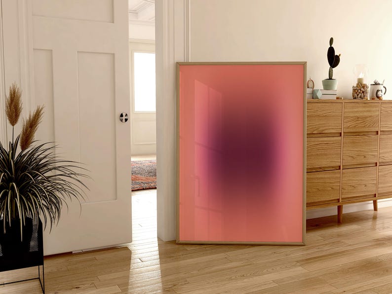 May include: A framed abstract art print with a pink and purple gradient. The print is hanging on a white wall in a room with wooden floors.