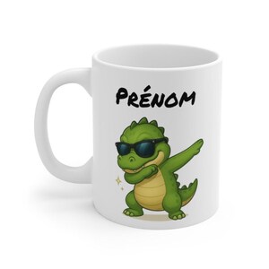 Peut inclure: Tasse en céramique blanche avec un dinosaure vert de dessin animé portant des lunettes de soleil et faisant un dab. Le mot "PRÉNOM" est imprimé en noir au-dessus du dinosaure.