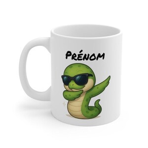 Peut inclure: Mug en céramique blanche avec un serpent vert de dessin animé portant des lunettes de soleil et faisant un dab. Le mot "PRÉNOM" est imprimé au-dessus du serpent. La tasse a une anse incurvée.
