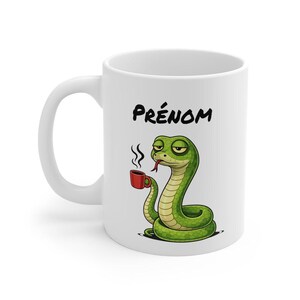 Peut inclure: Mug en céramique blanche avec un serpent vert de dessin animé tenant une tasse de café rouge. Le mug porte le mot "PRÉNOM" en lettres noires. Le serpent a une expression endormie, suggérant qu'il a besoin de plus de café.