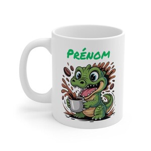 Peut inclure: Mug en céramique blanche avec un dessin d'alligator de dessin animé. L'alligator vert tient une tasse de café, entouré d'éclaboussures et de grains de café. Le mot "PRÉNOM" est imprimé en vert au-dessus de l'alligator.