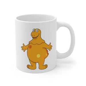 Peut inclure: Mug en céramique blanche avec un personnage de dessin animé orange avec des taches jaunes et un visage souriant. Le personnage a les bras tendus. La tasse a une anse incurvée.