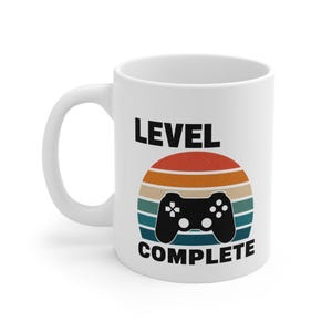 Peut inclure: Un mug en céramique blanche avec le texte noir LEVEL COMPLETE et un graphique de manette de jeu noire centré sur un motif de coucher de soleil semi-circulaire avec des rayures horizontales dans des tons rouge, orange, jaune et bleu sarcelle.