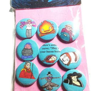May include: Eight round blue and white enamel pins featuring characters from the animated film Howl's Moving Castle. The pins are packaged on a pink background with a small image of the film's main characters in the top left corner. The text "Here's another curse; "May your bacon burn" is printed on the pink background.