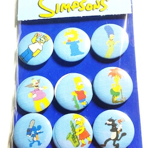 May include: A set of nine The Simpsons character buttons. The buttons are round and have a blue background with white outlines. Each button features a different Simpsons character, including Homer, Marge, Bart, Lisa, Maggie, Krusty the Clown, and Itchy and Scratchy.